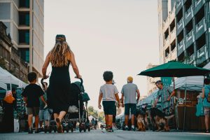 Family walking with stroller and dogs through lively outdoor market street on a sunny day.